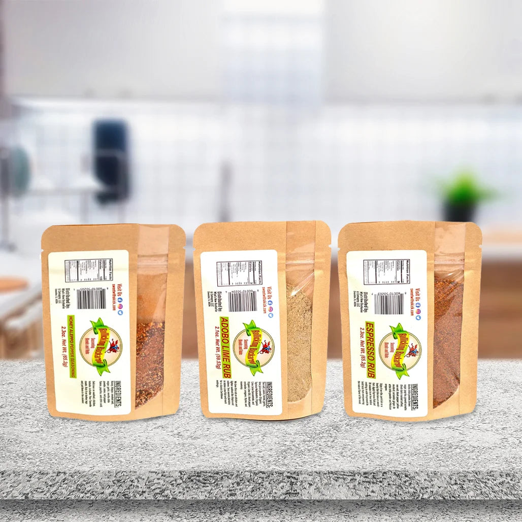 espresso rub, honey aleppo pepper seasoning, and adobe lime rub seasoning blends next to each other on a counter top and barbecue pit in the back